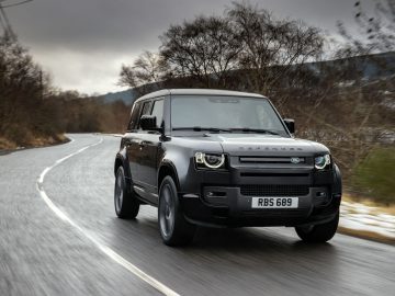 Een zwarte Land Rover Defender V8 met kenteken "RBS 689" rijdt op een bochtige weg geflankeerd door bladerloze bomen en stukken sneeuw.