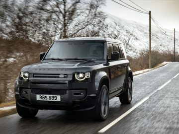 Een strakke zwarte Land Rover Defender V8 rijdt over een verharde weg met besneeuwde bomen en heuvels op de achtergrond.