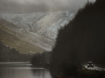 Een Land Rover Defender V8 rijdt op een smalle weg langs een kalm meer, met besneeuwde bergen en een donkere, beboste heuvel op de achtergrond onder een bewolkte hemel.