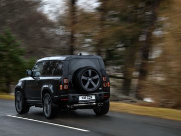 Een zwarte Land Rover Defender V8 rijdt op een natte weg omzoomd door vage bomen, met het reservewiel aan de achterkant van het voertuig gemonteerd.