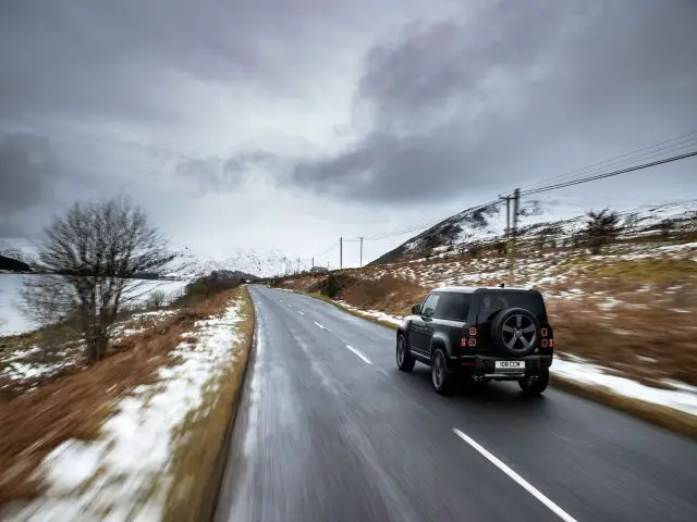 Een zwarte Land Rover Defender V8 rijdt op een natte, lege weg door een besneeuwd landelijk landschap met bergen in de verte.