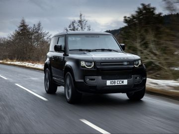 Bij bewolkt weer rijdt een zwarte Land Rover Defender V8 over een tweebaansweg omgeven door bomen.