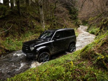 Een zwarte Land Rover Defender V8 rijdt door een ondiepe, rotsachtige stroom in een bosrijke omgeving. De omgeving is weelderig met groene vegetatie en bomen.