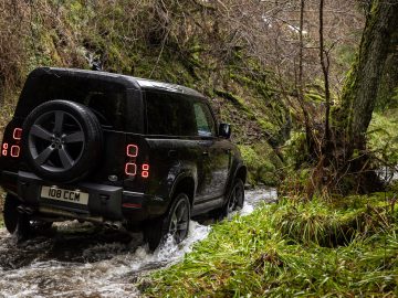 Een zwarte Land Rover Defender V8 steekt een ondiepe stroom over in een bosrijk gebied met met mos bedekte bomen en planten, en navigeert moeiteloos door het ruige terrein.