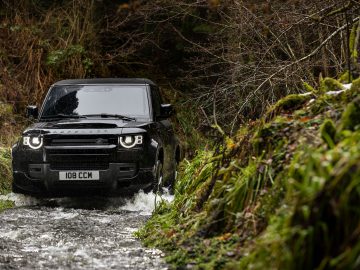 Een Land Rover Defender V8 rijdt door een ondiepe, rotsachtige stroom omgeven door weelderig groen gebladerte en bomen.