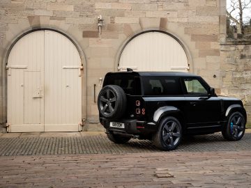 Een zwarte Land Rover Defender V8 staat geparkeerd op een geplaveid pad vlakbij een oud stenen gebouw met grote, gesloten houten deuren.