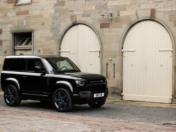 Een zwarte Land Rover Defender V8 staat geparkeerd voor een historisch stenen gebouw met twee grote, gesloten houten deuren.