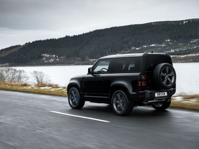 Een Land Rover Defender V8 rijdt op een weg langs een meer en beboste heuvels.