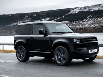 Een zwarte Land Rover Defender V8 SUV rijdt op een weg met een meer en beboste heuvels op de achtergrond.