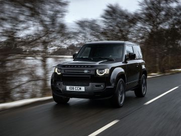 Een zwarte Land Rover Defender V8 rijdt op een weg met vage bomen en een watermassa op de achtergrond. Op het kenteken van het voertuig staat "108 CCM.