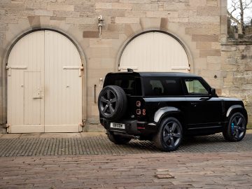 Een zwarte Land Rover Defender V8 staat geparkeerd op een geplaveide oprit voor een stenen gebouw met twee grote, gesloten houten garagedeuren.