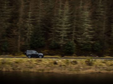 Een zwarte Land Rover Defender V8 rijdt over een smalle weg met een dicht bos en een watermassa op de achtergrond.