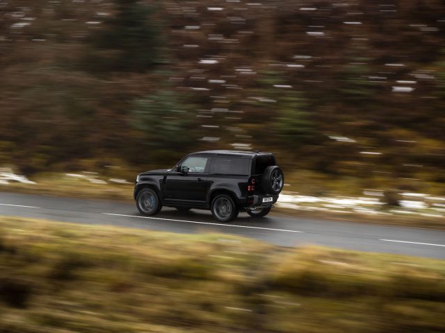 Een zwarte Land Rover Defender V8 rijdt op een landweg met een wazige bosachtergrond. Het voertuig rijdt naar de linkerkant van de afbeelding.