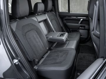 Het interieur van de Land Rover Defender V8 toont de achterbank met zwart lederen bekleding. In de middenstoel is een armleuning met bekerhouders neergeklapt, wat zowel luxe als gemak biedt.