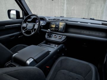 Binnenaanzicht van een Land Rover Defender V8 met het stuur, het dashboard met digitaal display en de middenconsole met bedieningsknoppen en versnellingspook. Aan de passagierszijde is een "Defender"-logo weergegeven.