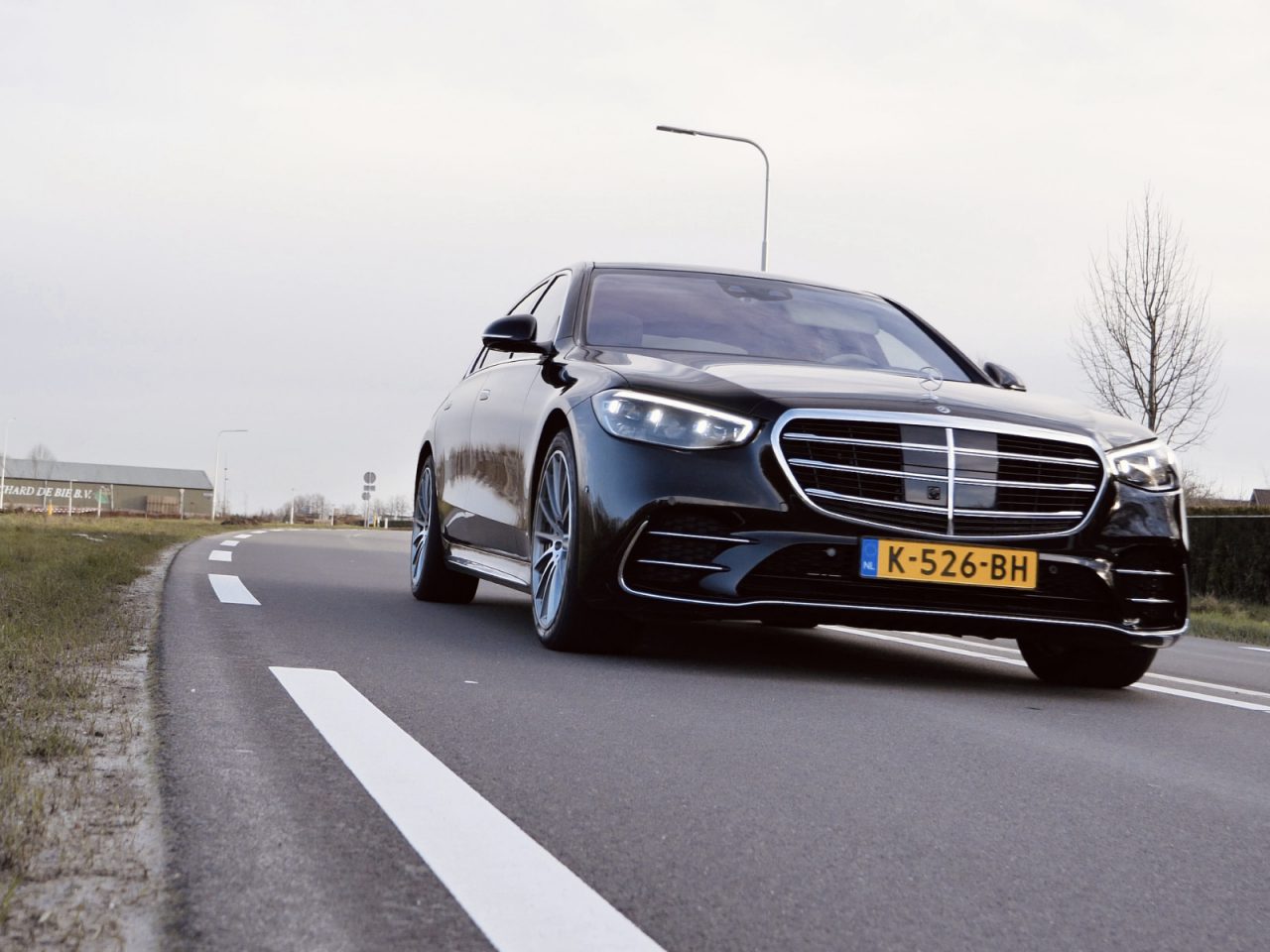 Een zwarte Mercedes-Benz S-Klasse sedan met kenteken "K-526-BH" rijdt op een tweebaansweg naast een grasveld en bomen, onder een bewolkte hemel.