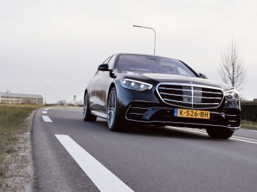 Een zwarte Mercedes-Benz S-Klasse sedan met kenteken "K-526-BH" rijdt op een tweebaansweg naast een grasveld en bomen, onder een bewolkte hemel.