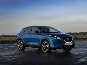 Een blauwe Nissan Qashqai SUV staat geparkeerd op een natte stoep, omlijst door een grasveld en een heldere lucht op de achtergrond.