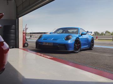 Een blauwe Porsche 911 GT3 staat geparkeerd bij de ingang van een racebaangarage naast een rode auto. In de garage is een stapel banden zichtbaar.