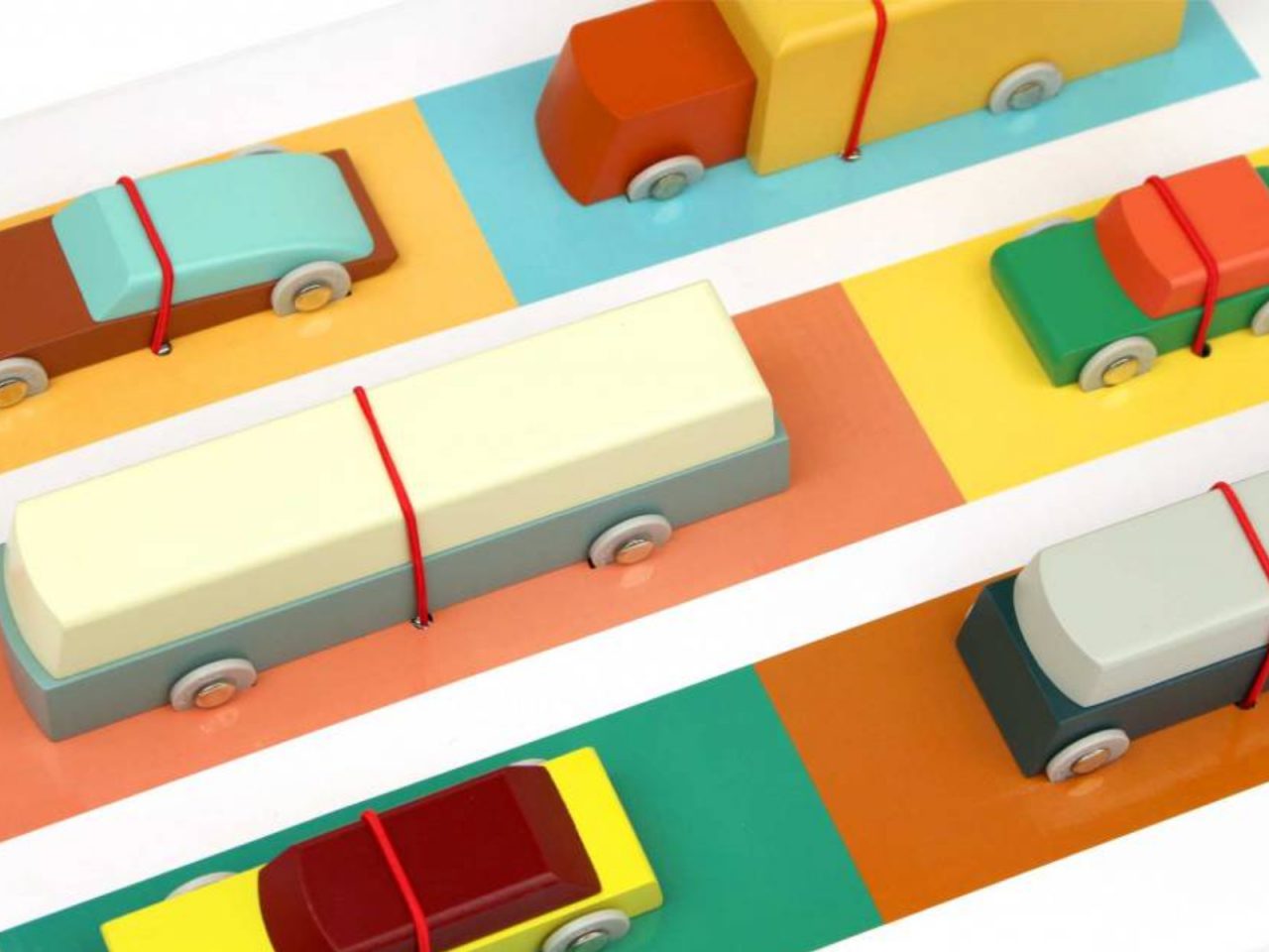 Kleurrijke speelgoedvoertuigen, die doen denken aan de ontwerpen van Floris Hovers, worden vastgezet met rode touwtjes en op een veelkleurige mat met bijbehorende kleurblokken geplaatst.