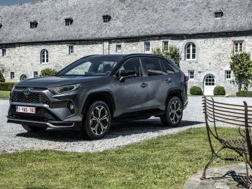 Een grijze Toyota RAV4 Plug-in Hybrid staat geparkeerd op een grindoprit voor een stenen gebouw met een leien dak, omgeven door gras en een houten bank op de voorgrond.