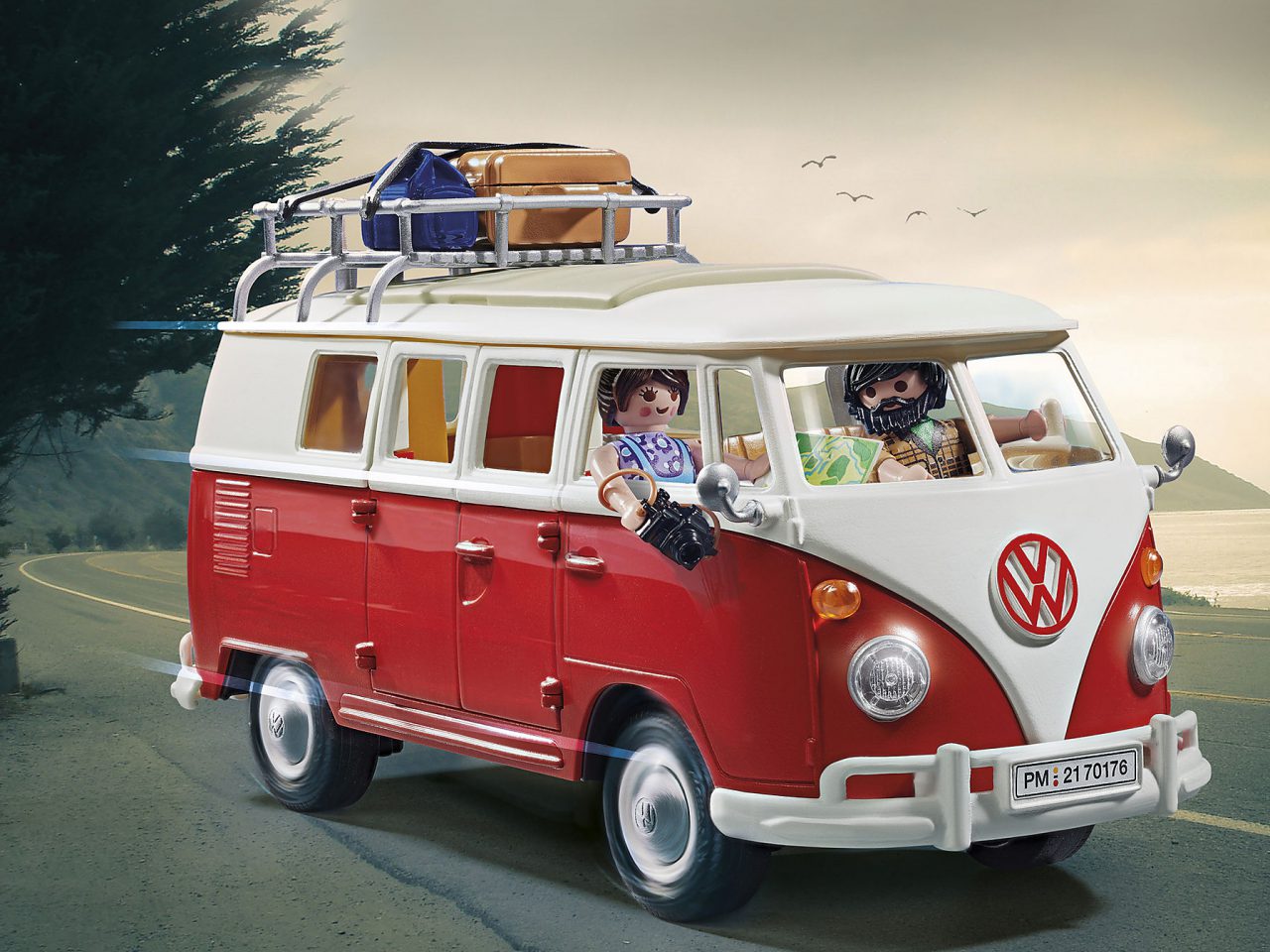 Een rood-wit model VW-camper met Playmobil-passagiers rijdt op een kustweg. Het busje heeft bagage op het dak en de lucht is bewolkt.