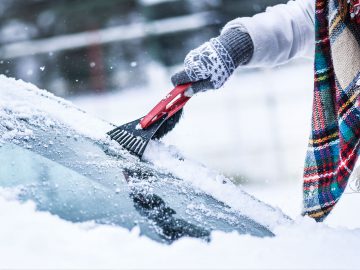 Persoon die handschoenen en een geruite sjaal draagt, gebruikt een ijskrabber om sneeuw van de voorruit van een auto te verwijderen op een ijskoude, besneeuwde dag.