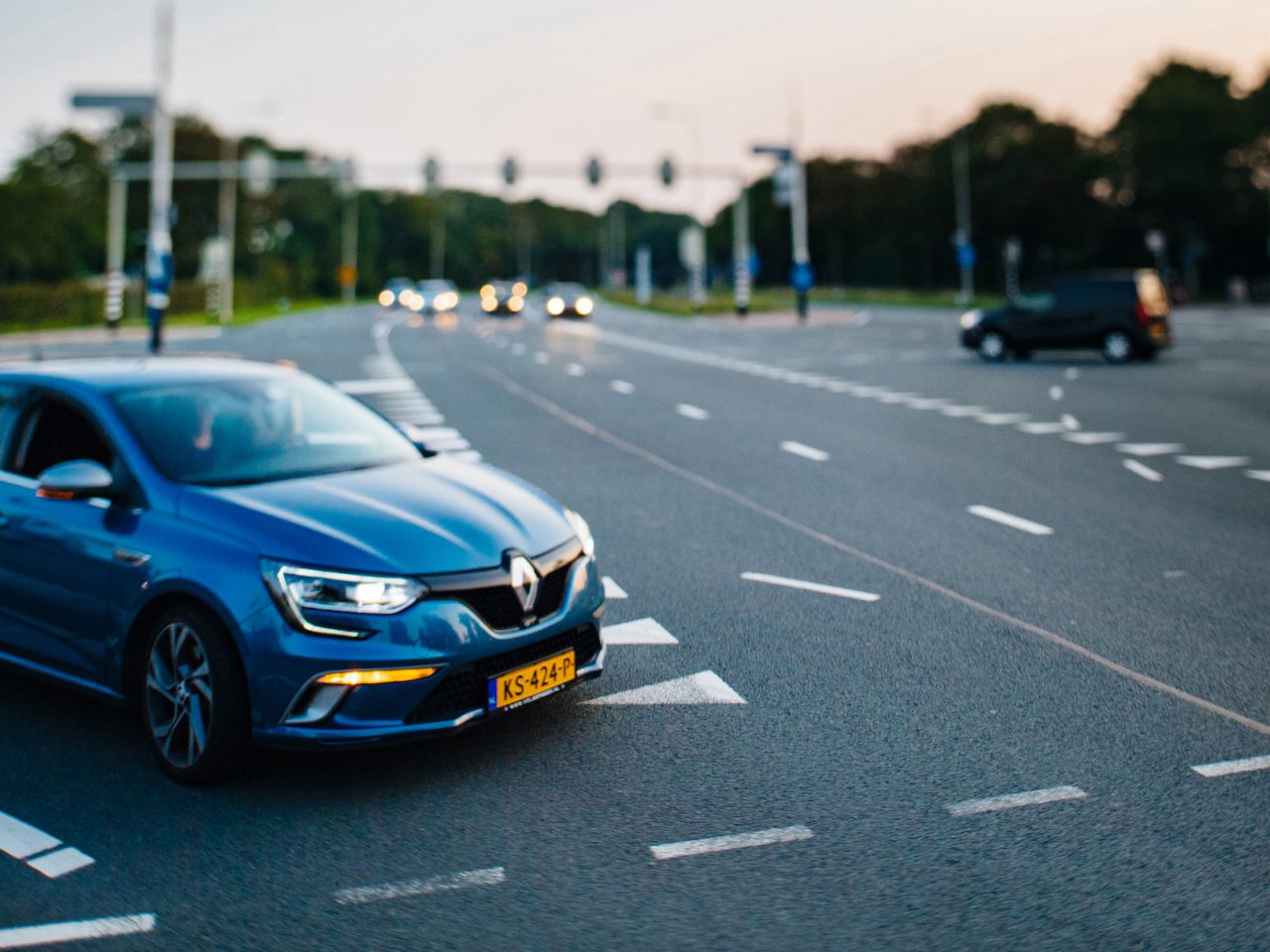 Een blauwe auto met een Nederlands kenteken, bestuurd door iemand die waarschijnlijk opgewonden is omdat hij onlangs zijn autorijbewijs heeft gehaald, maakt een bocht op een kruispunt met meerdere rijstroken, verkeerslichten en een paar andere voertuigen op de achtergrond onder een schemerige avondlucht.