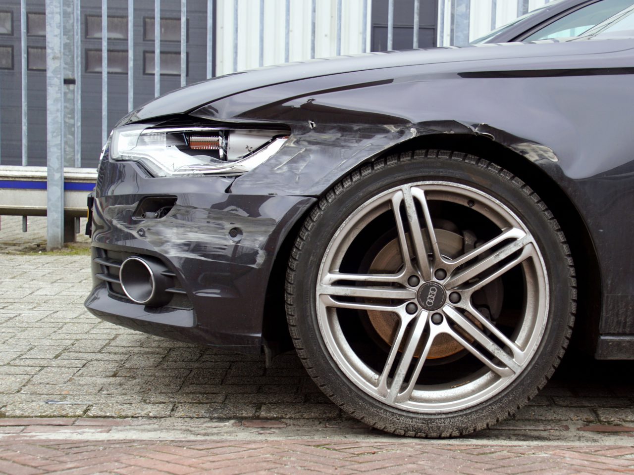 Een zwarte auto met zichtbare "minder autoschade" aan het linker voorspatbord en de bumper, geparkeerd op een verharde ondergrond nabij een metalen hek.
