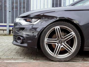 Een zwarte auto met zichtbare "minder autoschade" aan het linker voorspatbord en de bumper, geparkeerd op een verharde ondergrond nabij een metalen hek.
