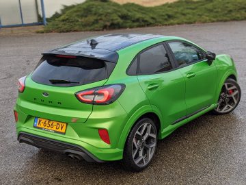 Een groene Ford Puma ST compacte crossover staat geparkeerd op een natte weg in de buurt van weelderige groene struiken. Hij beschikt over een achterspoiler, dubbele uitlaat en het kenteken "K-656-DV.