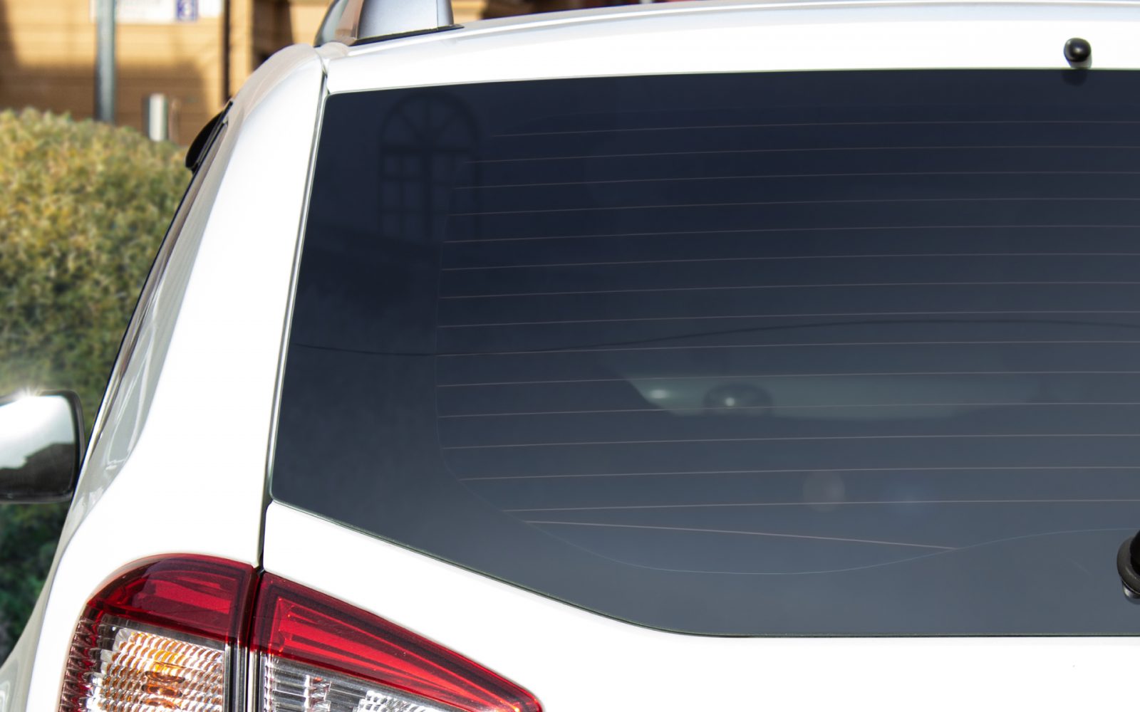 Getoond worden de achterruit en het achterlicht van een wit voertuig met geblindeerde autoruiten. De achtergrond omvat wat gebladerte en een deel van een gebouw.