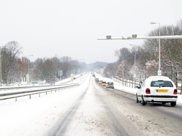 Een besneeuwde weg met meerdere auto's die in beide richtingen rijden onder toeziend oog van een verkeerscontrole- of tolportaal. Winterweer heeft de zijkanten van de weg en de bomen bedekt, waardoor een schilderachtig wintertafereel ontstaat.