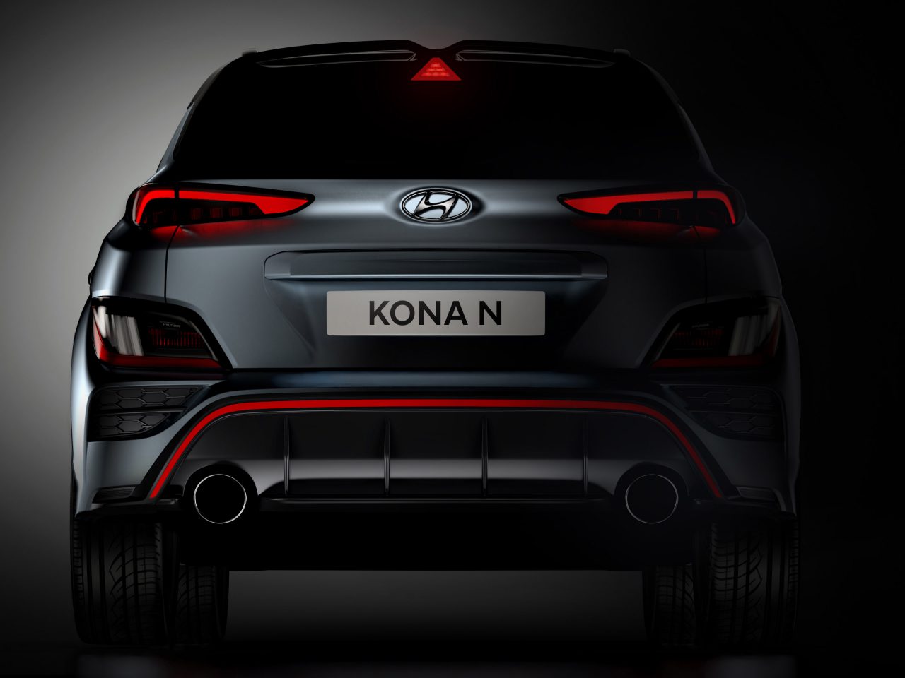 Achteraanzicht van een donkergekleurde Hyundai Kona N SUV met verlichte rode achterlichten, een zichtbaar Hyundai-logo en dubbele uitlaten, tegen een donkere achtergrond.