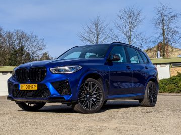 Een blauwe BMW X5 SUV staat geparkeerd op een verhard terrein onder een heldere hemel, met kale bomen en een laag gebouw op de achtergrond. Het voertuig heeft een geel kenteken met het nummer "H-100-RP.