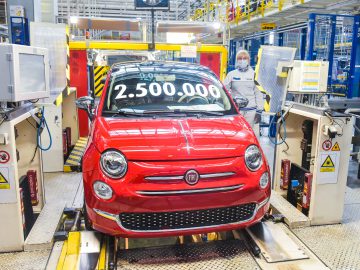 Een rode Fiat 500-auto met "2.500.000" op de voorruit staat op een lopende band in een autofabriek. Ernaast staat een arbeider, met een masker en een wit uniform op.