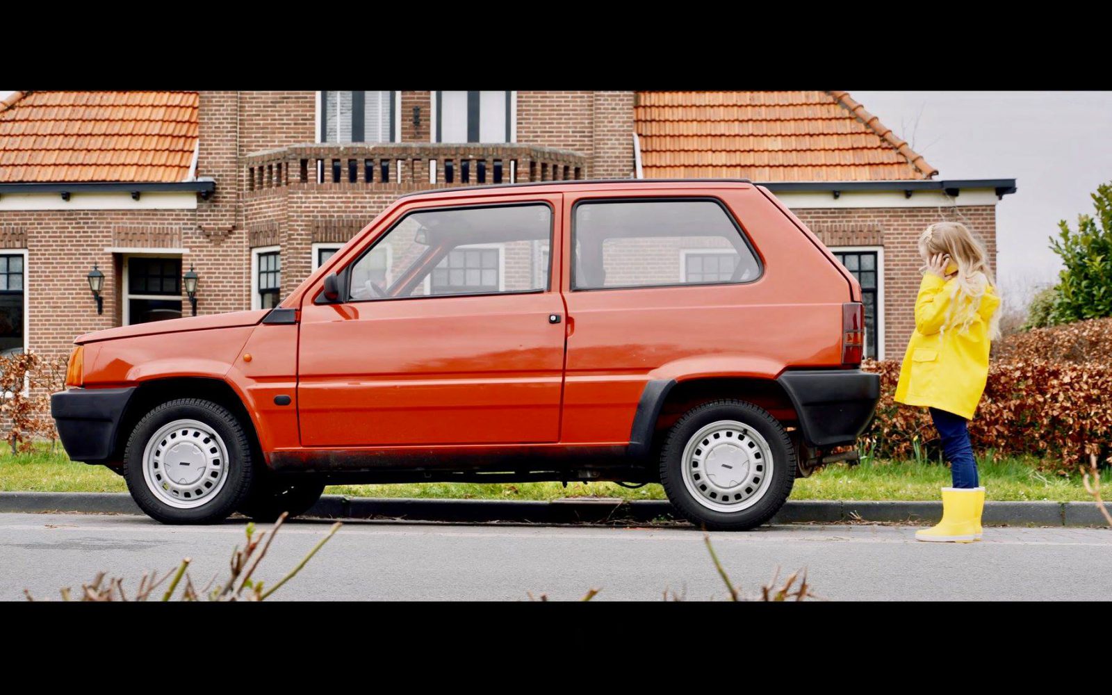 Een kind in een gele regenjas en laarzen staat naast een rode Fiat Panda die op straat geparkeerd staat, met een bakstenen huis op de achtergrond.