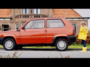 Een kind in een gele regenjas en laarzen staat naast een rode Fiat Panda die op straat geparkeerd staat, met een bakstenen huis op de achtergrond.