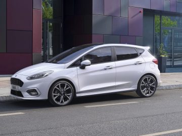 Een witte hatchback, waarschijnlijk afkomstig van het evenement autoverkoop februari 2021, staat geparkeerd aan de kant van een straat voor een gebouw met paarse en kastanjebruine panelen.