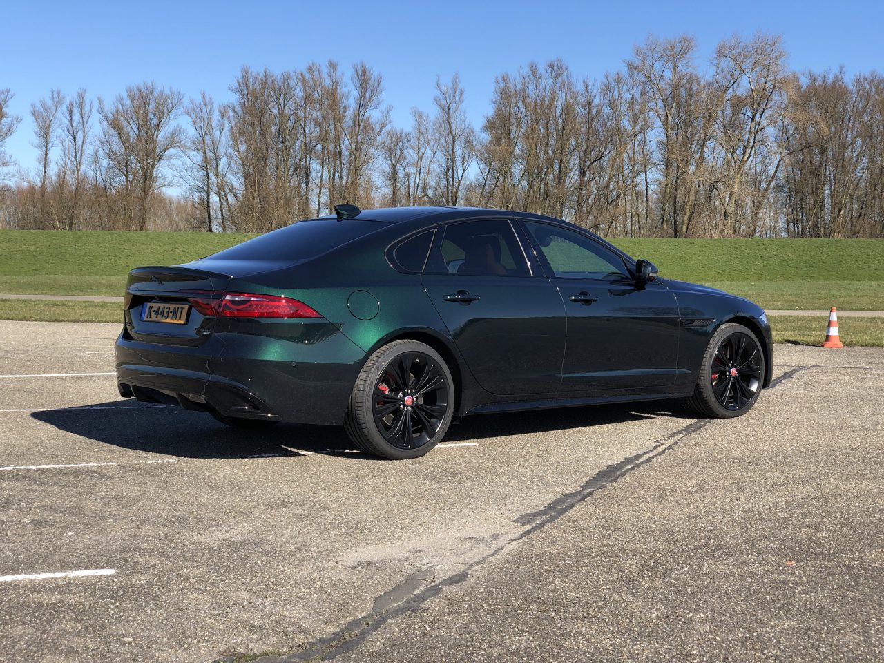 Een donkergroene Jaguar XF sedan staat geparkeerd op een lege parkeerplaats met een grasveld en bladerloze bomen op de achtergrond onder een helderblauwe lucht.