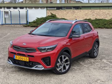 Een rode Kia Stonic compacte SUV met Nederlands kenteken, geparkeerd op een verhard oppervlak buiten, voor een gebouw met een bakstenen gevel.