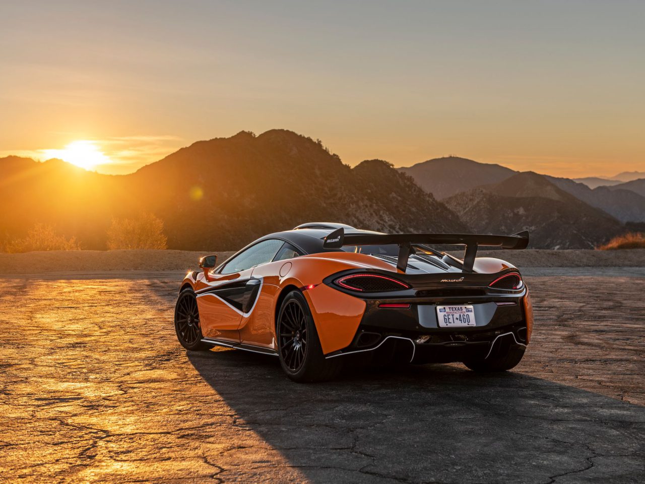 Een feloranje McLaren-sportwagen met een spoiler staat geparkeerd op een schilderachtige berg met uitzicht op zonsondergang. De zon gaat achter de heuvels onder en werpt een oranje gloed over het landschap.