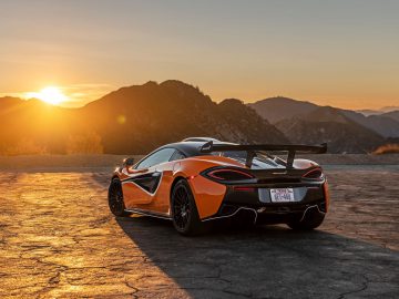 Een feloranje McLaren-sportwagen met een spoiler staat geparkeerd op een schilderachtige berg met uitzicht op zonsondergang. De zon gaat achter de heuvels onder en werpt een oranje gloed over het landschap.