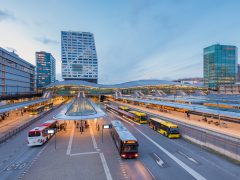 Een modern busstation met diverse bussen en enkele passagiers, omgeven door hoge gebouwen aan het begin van de avond, toont de impact van de initiatieven van Mobiliteitsalliantie op het gebied van het verbeteren van het stadsvervoer.