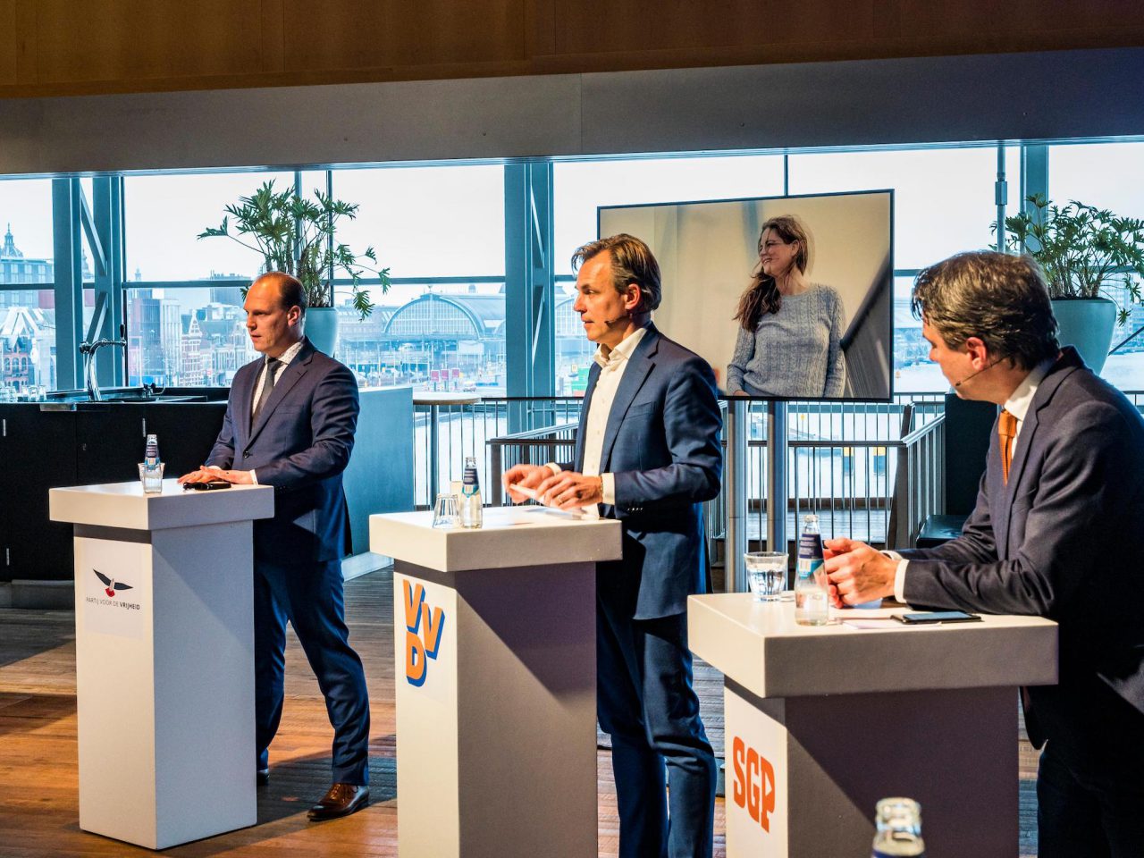 Drie mannen in zakenkleding staan op podia met respectievelijk de opschriften CU, VVD en SP en nemen deel aan het Mobiliteitsdebat. Op de achtergrond is een vrouw zichtbaar op een scherm. De scène speelt zich binnenshuis af met grote ramen.