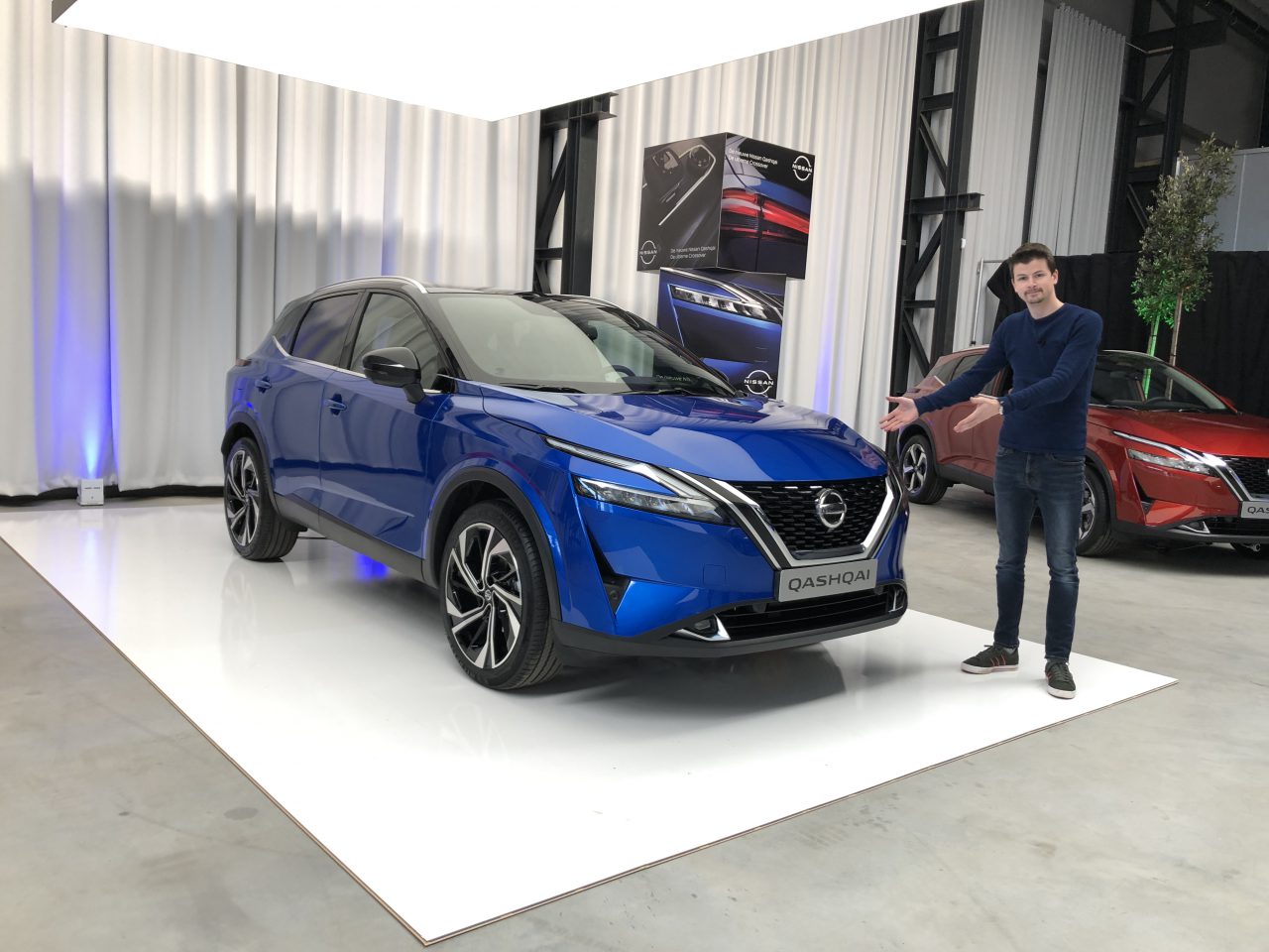 Een persoon staat naast een blauwe Nieuwe Nissan Qashqai op een wit platform, met op de achtergrond een andere rode auto zichtbaar.