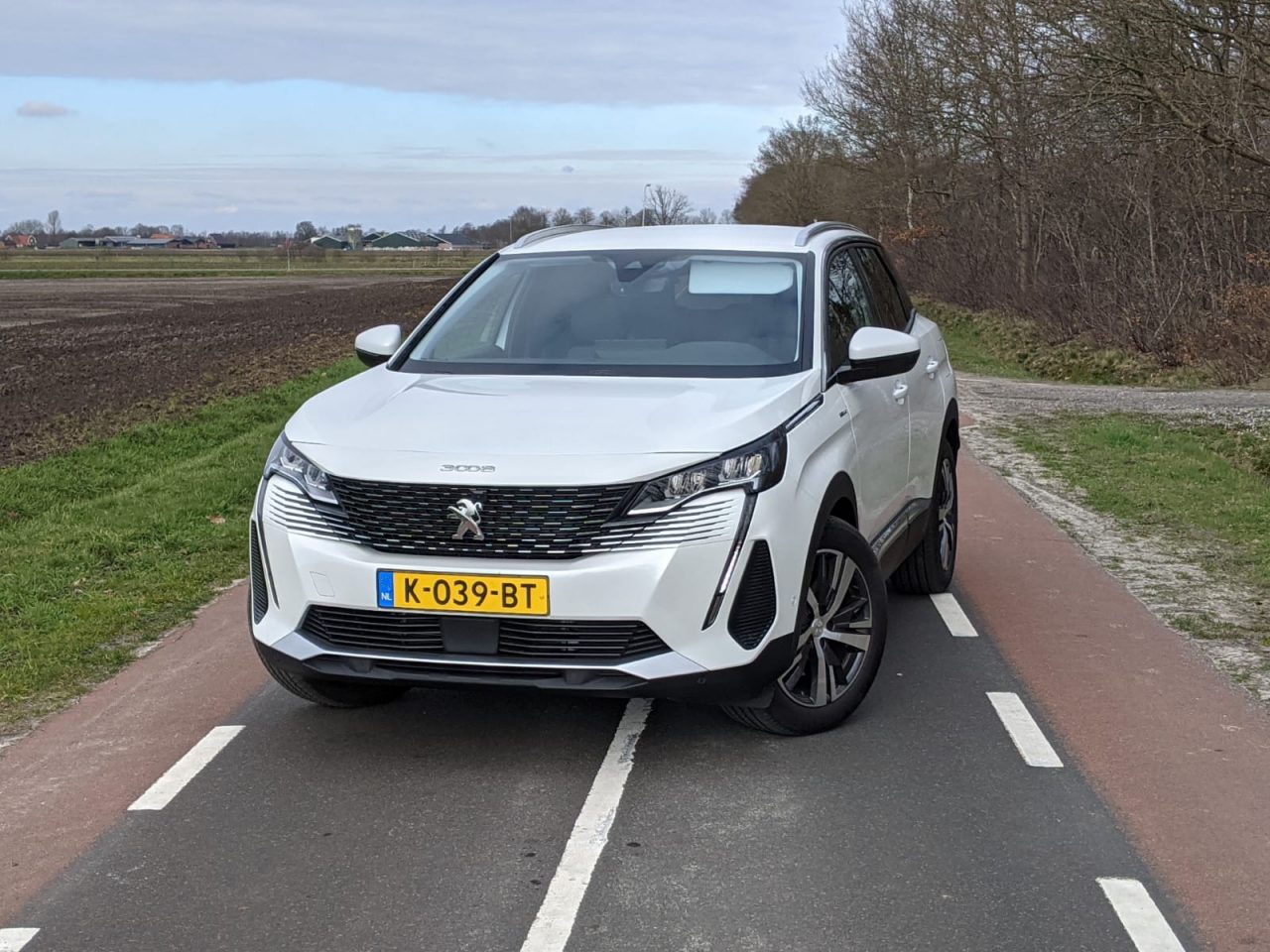 Een witte Peugeot 3008 SUV staat geparkeerd op een weg met aan de ene kant velden en aan de andere kant bomen. Op het kenteken staat "K-039-BT".