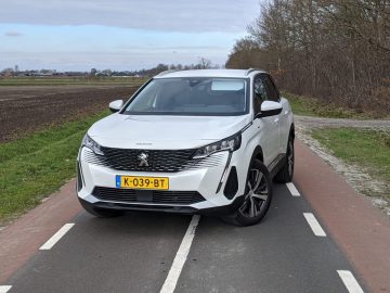 Een witte Peugeot 3008 SUV staat geparkeerd op een weg met aan de ene kant velden en aan de andere kant bomen. Op het kenteken staat "K-039-BT".