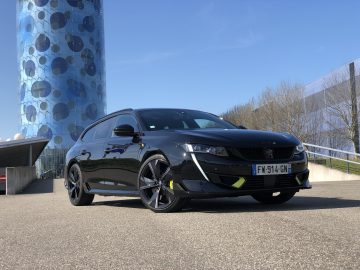 De strakke Peugeot 508 PSE sportwagen staat geparkeerd op een verharde ondergrond, met een modern glazen gebouw en een helderblauwe lucht op de achtergrond.