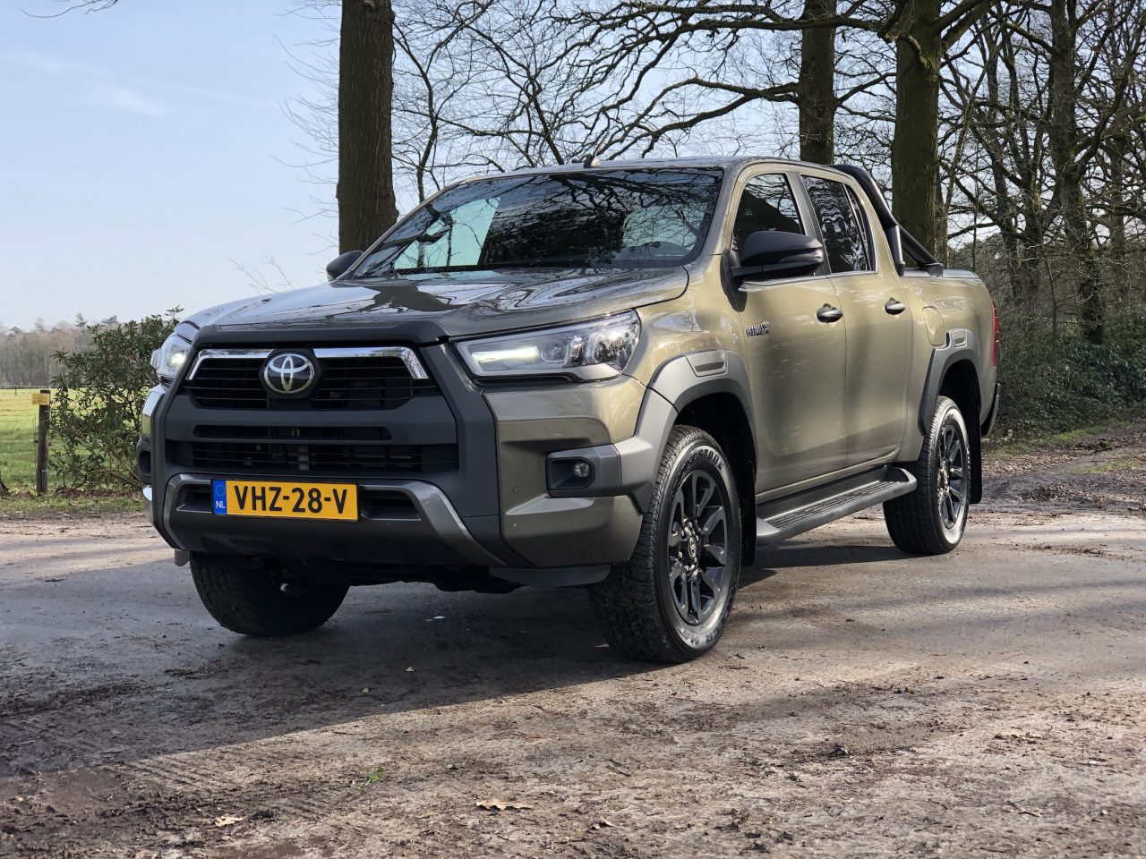 Een groene Toyota Hilux pick-up staat geparkeerd op een onverharde weg met bomen op de achtergrond. Het voertuig heeft een Nederlands kenteken, VHZ-28-V.